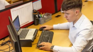 foto de um jovem branco, cabelos castanhos, vestindo uma camisa de manga comprida branca, em um ambiente de trabalho, sentado em uma estação de trabalho de madeira clara, na frente de um computador e um notebook, olhando para a tela do computador, segurando o mouse com a mão direita e com a outra apoiada no teclado. Ao fundo, uma garrafinha preta térmica e porta trecos com lápis e canetas.