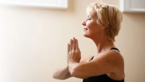 foto de uma senhora loira, de cerca de 70 anos, vestindo camiseta preta e com as mãos unidas na altura do peito em uma posição meditativa típica de ioga. A imagem só mostra seu perfil até a altura do peito.