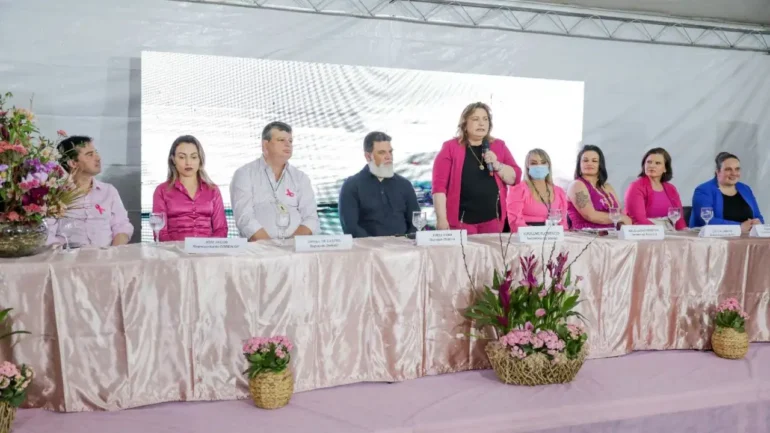 foto de 5 mulhers vestidas de rosa, uma outra vestida de azul, um homem com camisa rosa, outro de branco e outro de preto sentados a frente de uma grande mesa com toalha branca, em cima de um palco, em uma cerimônia de abertura de um evento de Reconstrução Mamária.