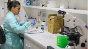 foto de uma mulher em um laboratório de análise clínica vestindo roupão de proteção verde claro, luvas azuis, cercada por vários produtos e instrumentos laboratoriais.