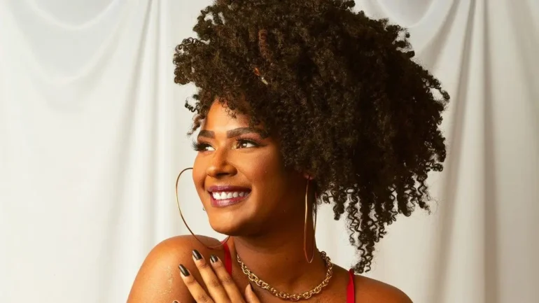 foto do rosto de uma bonita mulher negra, com os cabelos cacheados amarrados para cima, brincos enormes de argola e colar, sorrindo e olhando para frente.