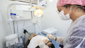 foto de uma dentista atendendo uma paciente mulher. A dentista está com máscara, óculos de graus, toca de pano rosa prendendo os cabelos, luvas e está examinando os dentes da cliente com um espelhinho caneta. A paciente está com calá xadrez preta e branca, sapatos pretos, blusa preta, toalha sobre o peito, óculos de proteção e aboca aberta.