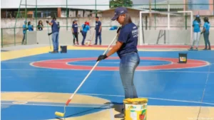 foto de uma jovem com boné azul, camiseta azul e calça jeans, pitando o chão de uma quadra de esporte. Ao fundo, outros 9 jovens também com camisetas azuis também participam.