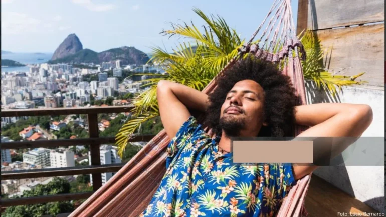 foto de um homem mulato deitado na rede com os olhos fechados e um sorriso no rosto, vestindo blusa estampada azul, ao fundo a imagem da cidade do Rio de Janeiro.