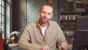 foto de um homem com os cabelos castanhos claros amarrados ao alto da cabeça, com barba, vestindo uma camieste branca e um pullover marrom claro por cima, sentado em frente a uma mesa com um computador a frente, com um leve sorriso no rosto e olhando para a câmera. Ao fundo, uma estante com copos e uma máquina de café e um armário de ferro.