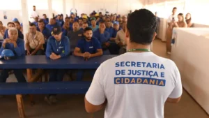 Foto de um homen de costas olhando para um público de vários empregados uniformizados com camisetas azuis, de uma obra, sentados em bancos em frente a mesas de madeira azuis olhando para ele.