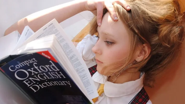 foto de uma menina loira, encostada em um sofá, vestindo uniforme escola de camisa branca, gravata amarela, e suspensório quadriculada com tons vermelho escuro, lendo um diciona´rio de inglês da Oxford.