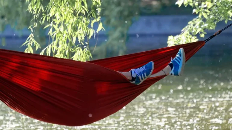 foto de uma rede vermelha bem viva, com uma pessoa deitada com os 2 pés calçando tênis azul para fora da rede, Ao fundo, folhas da árvore e um rio.