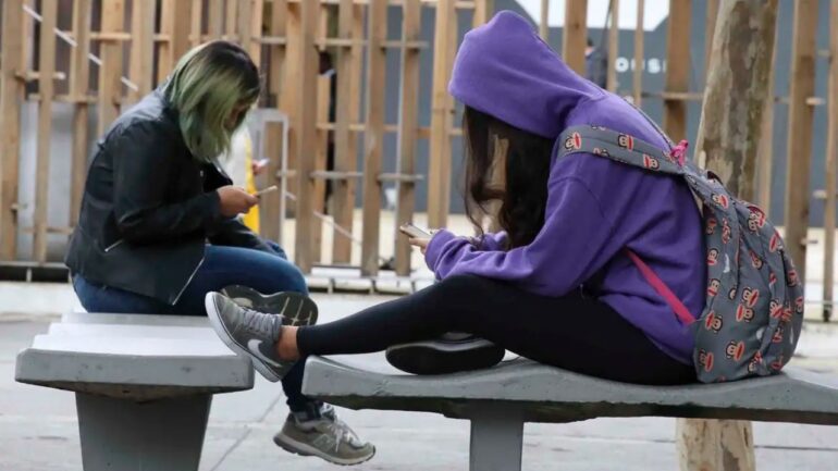 foto de duas estudantes sentadas cada uma em um banco de cimento de uma escola pública olhando cada uma para o seu celular. A menina da direita veste calças jeans, tênis e um casaco de capuz roxo. A outra, jeans casaco preto e tênis. Ao fundo, a grade amarela da cerca da escola.