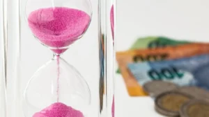 imagem de uma ampulheta de vidro transparente com areia rosa caindo e ao fundo desfocadas várias notas de dinheiro e moedas.