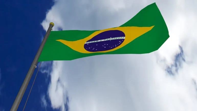 imagem da bandeira do Brasil vista de baixo para cima, presa em um mastro. Ao fundo o céu cheio de nuvens.