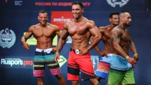 Foto de 4 fisioculturistas sorridentes, no palco, de concurso fazendo poses. Todos estão vestindo bermudas coloridas até a altura dos joelhos.