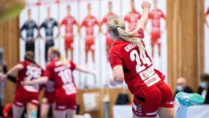 foto de uma jogadoras de handebol jogando. Em primeiro plano, uma jogadora loura está com o braço direito estendido para cima com o punho cerrado, comemorando e as demais estão ao fundo se cumprimentando. Elas estão em quadra, vestindo uniforme vermelho de camiseta e shorts. No fundo aparece um banner grande com a imagem delas e à direita, uma mesa lateral com 1 homem e 1 mulher com roupa executiva sentados e usando máscaras cirúrgicas.