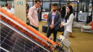 foto de 2 homens e 1 mulher conversando em um evento de tecnologia realizado em Brasília. A mulher tem cabelos castanhos escuros e veste conjunto branco de calça e blazer. Um dos homens veste camisa rosa clara e calça jeans, o outro homem calça justa, camisa e casaco. Os 3 olham para uma placa de captação de energia solar.