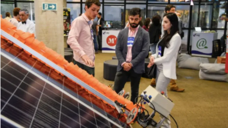 foto de 2 homens e 1 mulher conversando em um evento de tecnologia realizado em Brasília. A mulher tem cabelos castanhos escuros e veste conjunto branco de calça e blazer. Um dos homens veste camisa rosa clara e calça jeans, o outro homem calça justa, camisa e casaco. Os 3 olham para uma placa de captação de energia solar.