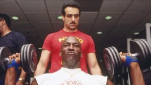 foto do jogador de basquete MICHAEL JORDAN sentado, vestindo uma camiseta branca com dizeres AIR JORDAN fazendo levantamento de peso, tendo seu treinador Tim Grover, vestindo uma camiseta vermelha com dizeres em amarelo, atrás olhando o movimento.