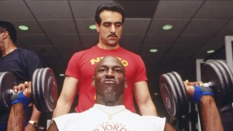 foto do jogador de basquete MICHAEL JORDAN sentado, vestindo uma camiseta branca com dizeres AIR JORDAN fazendo levantamento de peso, tendo seu treinador Tim Grover, vestindo uma camiseta vermelha com dizeres em amarelo, atrás olhando o movimento.