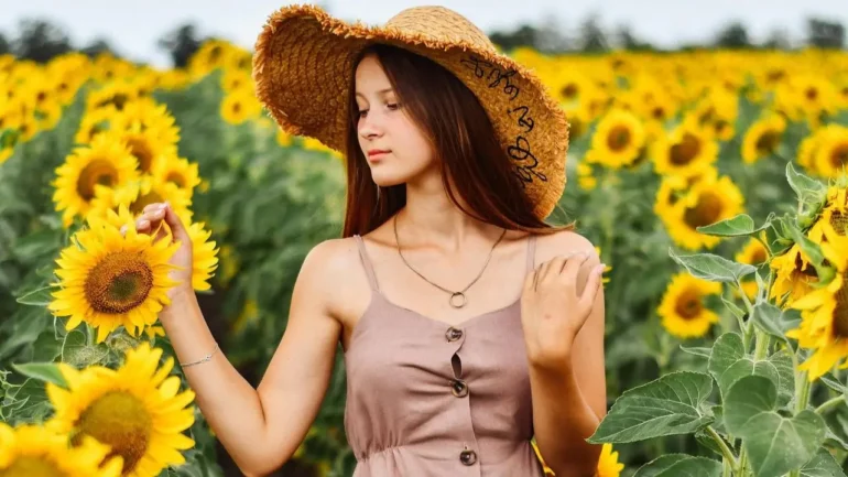 uma moça bonita de cabelos castanhos soltos, vestindo rande chapéu de palha, colar com uma aliança no pescoço, e um vestido marrom claro olhando para um girassol, no meio de uma plantação de girassóis.