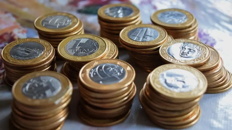 foto de várias pilhas de moedas de R$ 1,00 sobre um tampo de mesa de madeira.