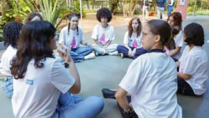 9 estudantes meninas sentadas no chão do pátio da escola, formando um círculo e conversando. Elas estão vestindo calças e camisetas de um programa Meninas em Ação do GDF.