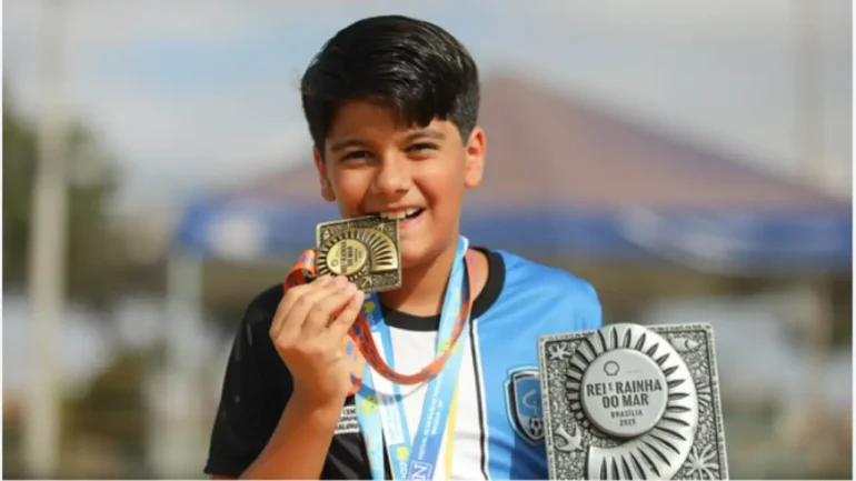 foto de um adolescente de cabelos castanhos escuros, vestindo blusa azul de uniforme e mordendo uma medalha de ouro, pendurada no pescoço, enquanto segura com a mão esquerda um troféu de natação.