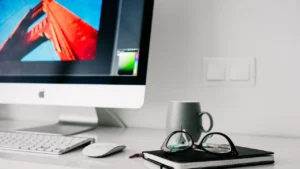 foto de parte de um computador IMAC, com uma imagem vermelha e azul, um mouse, perte de um teclado, um caneco, uma agenda preta um um óculos e grau de armação preta em cima da agenda. tudo isso em cima de uma mesa branca, que aparece apenas uma parte. O fundo é uma parede branca e o ambiente está claro, com a luz do dia.