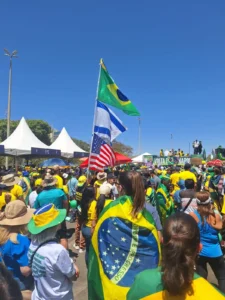 foto de manifestação popular com vários cidadãos vestidos de verde e amarelo e ao fundo a bandeira do Brasil, de Israel, e dos EUA flamejando. O céu azul, e o sol destacam o calor do dia 7 de setembro de 2025.