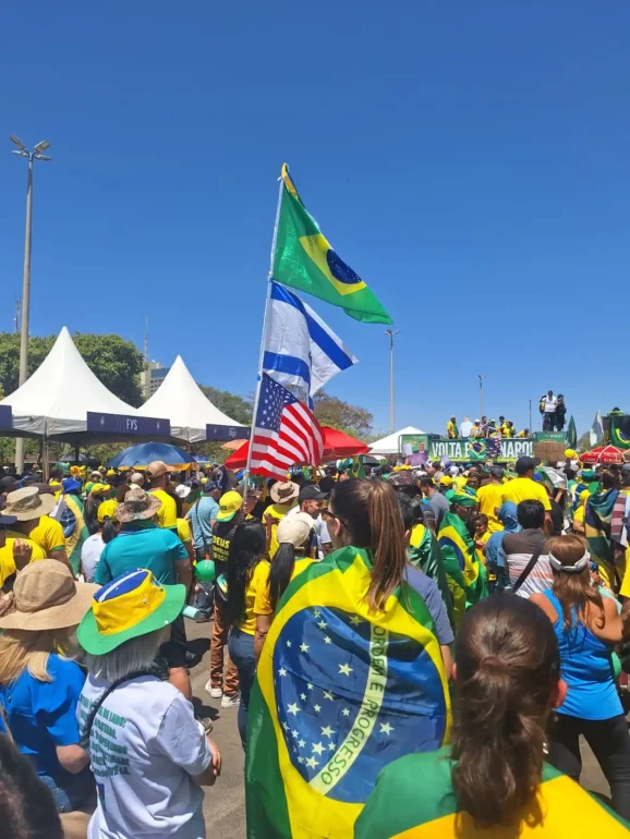 foto de manifestação popular com vários cidadãos vestidos de verde e amarelo e ao fundo a bandeira do Brasil, de Israel, e dos EUA flamejando. O céu azul, e o sol destacam o calor do dia 7 de setembro de 2025.
