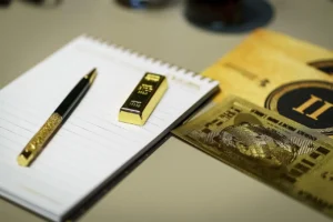 foto de uma barra de ouro e uma caneta de ouro e couro em cima de um bloco de notas de papel com espiral. Ao lado 2 placas finas de ouro, com imagens gravadas em relevo. Tudo sobre um tampão do que parece ser uma mesa de cor bege.