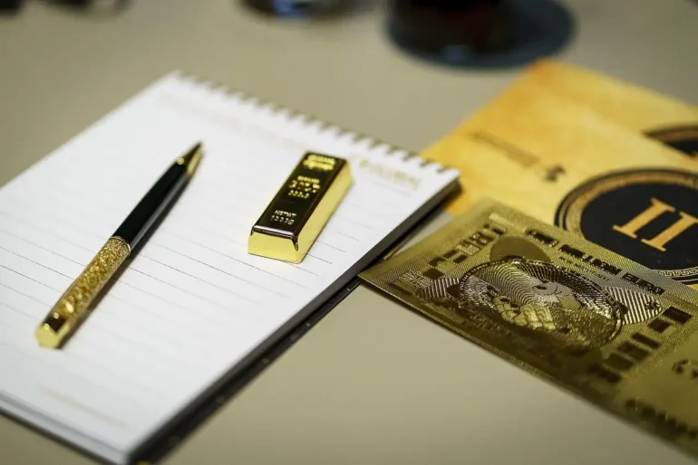 foto de uma barra de ouro e uma caneta de ouro e couro em cima de um bloco de notas de papel com espiral. Ao lado 2 placas finas de ouro, com imagens gravadas em relevo. Tudo sobre um tampão do que parece ser uma mesa de cor bege.