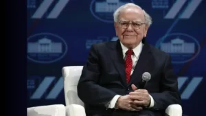 foto do homem mais rico do mundo WARREN BUFFET, sentado em uma poltrona pequena branca, vestindo um terno completo preto, camisa branca e gravata vermelha escura, com óculos e segurando nas duas mãos unidas um microfone, apoiado na perna. Ao fundo banners do evento em azul e branco.