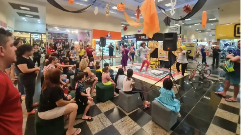 foto de centenas de adultos e crianças sentadas, dentro da praça central do shoping em frente a um palco, assistindo teatrinho do Detran de campanha educativa do trânsito.