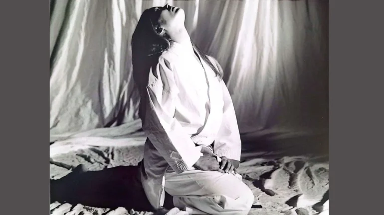 Foto preto e branco da carateca Carla Ribeiro vestida de quimono branco e faixa-preta, sentada em cima dos joelhos e alongando o pescoço para cima, com as mãos apoiadas no joelho. Ela está em cima de panos e ao fundo uma cortina branca cobre o cenário atrás dela.