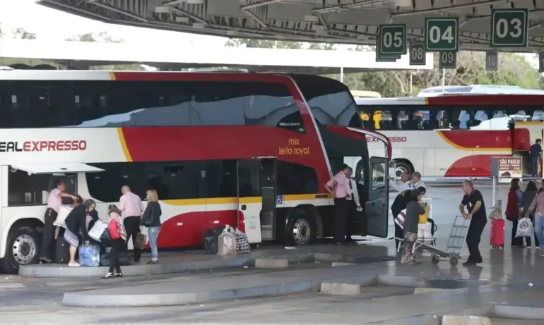foto de um ônibus leito vermelho, com detalhes brancos, de 2 andares estacionado na rodoviária de Brasília. Ao lado passageiros despacham malas para entrar no ônibus.