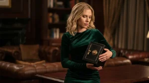 Imagem de uma loira parecida com a Scarlet Jonhson vestindo um vestido verde e olhando para um pequeno cofre em suas m&atilde;os. Ao fundo um escrit&oacute;rio cl&aacute;ssico com m&oacute;veis escuros.