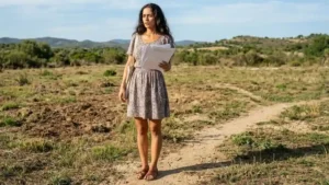 imagem de uma morena parecida com a atriz Sônia Braga, vestindo um vestidinho simples e estampada, com sandálias baixas de couro, segurando uns papéis e olhando o terreno a frente. Ao fundo um terreno, parecido com o cerrado
